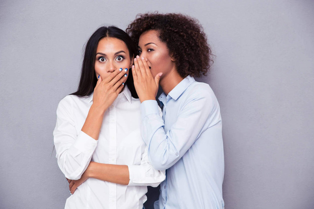 Two women sharing a secret, one covering her mouth in surprise while the other whispers.