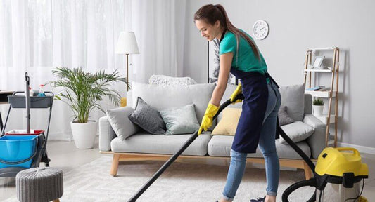 A house cleaner in a blue shirt and gloves vacuuming a modern living room with a sofa and plants.